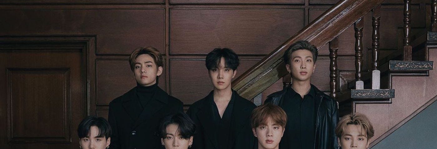 This is a group portrait in front of a straight wooden staircase. Top: V, j-hope, RM. Bottom: Suga, Jungkook, Jin, Jimin.