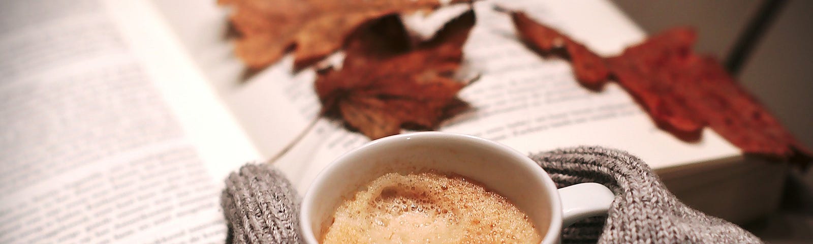 A cup of coffee, with a pair of grey gloves, an open book and some autumn leaves sitting on the pages.