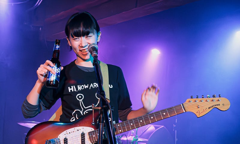 Photo of young woman onstage with an electric guitar and a beer on a violet blue background.