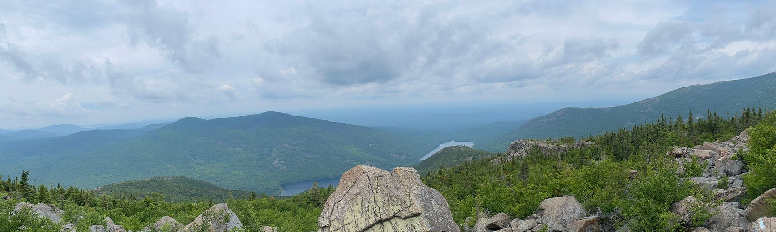 Mountaintop view with large boulders in the foreground and green mountain peaks in the background. In the middle, below, you can see a small lake peeking out. The sky is hazy with clouds and sun.