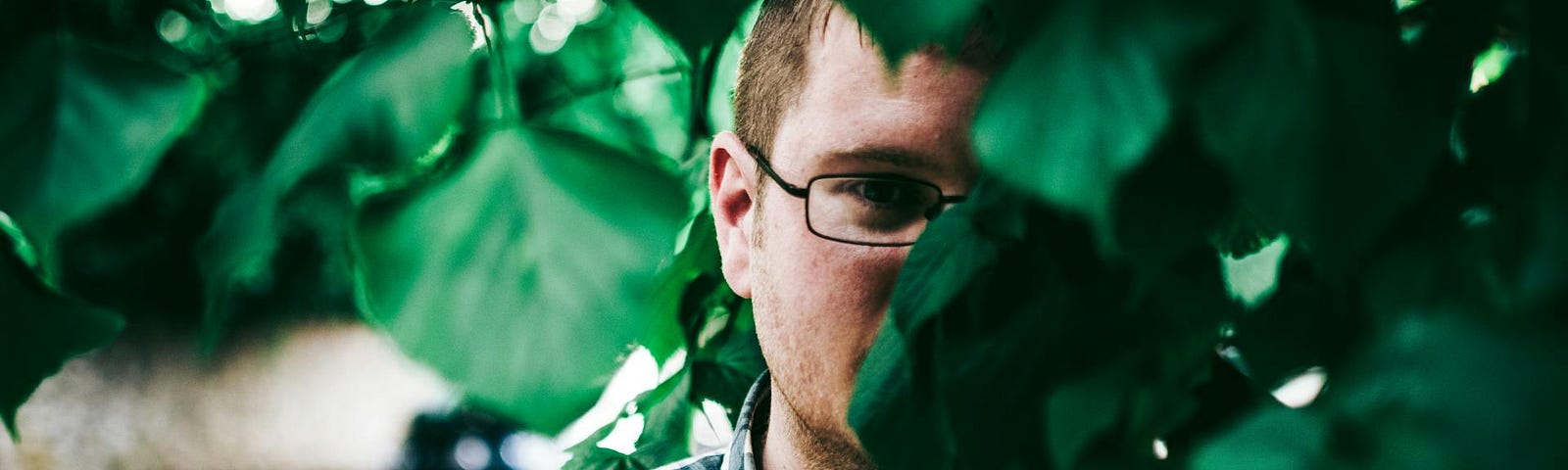 A man with eyeglasses standing behind green leaves.