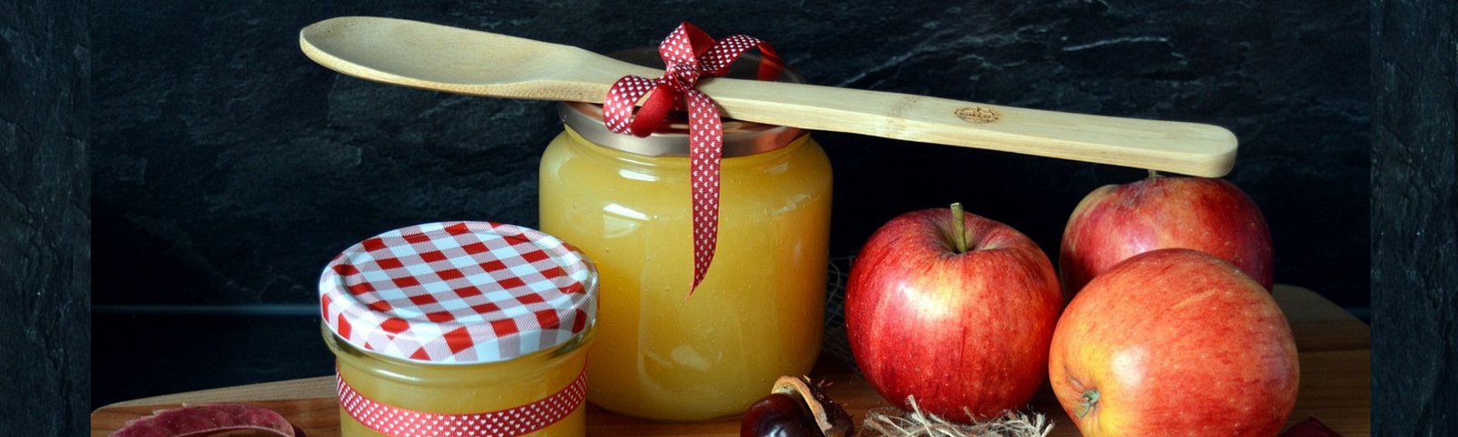 The makings of applesauce are arranged neatly on a wooden cutting board, including reddish-yellow apples, jars of chunky yellow sauce, a wooden spoon, with fall decor details like gingham ribbons around the spoon and one jar and as a pattern on the jar’s lid, two little wooden heart cut-outs, and a scrap of burlap.