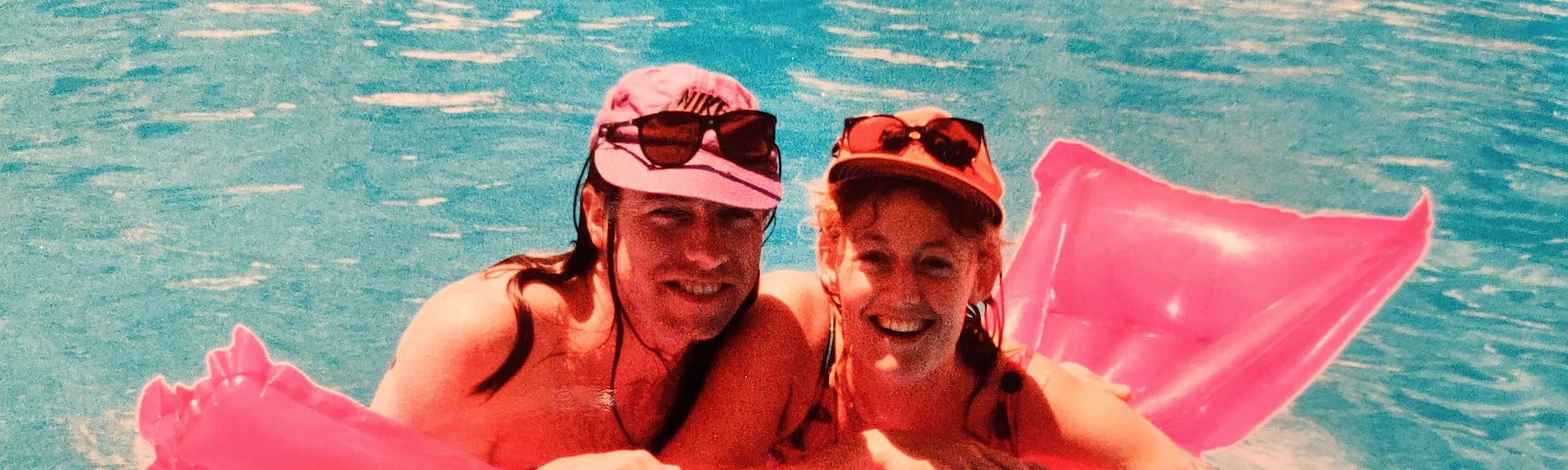 Two adults holding onto a bright pink inflatable airbed in a swimming pool in summer.