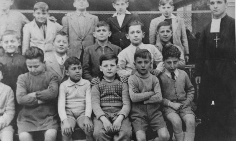 European, and mostly Jewish, children, sit with a priest during World War II.