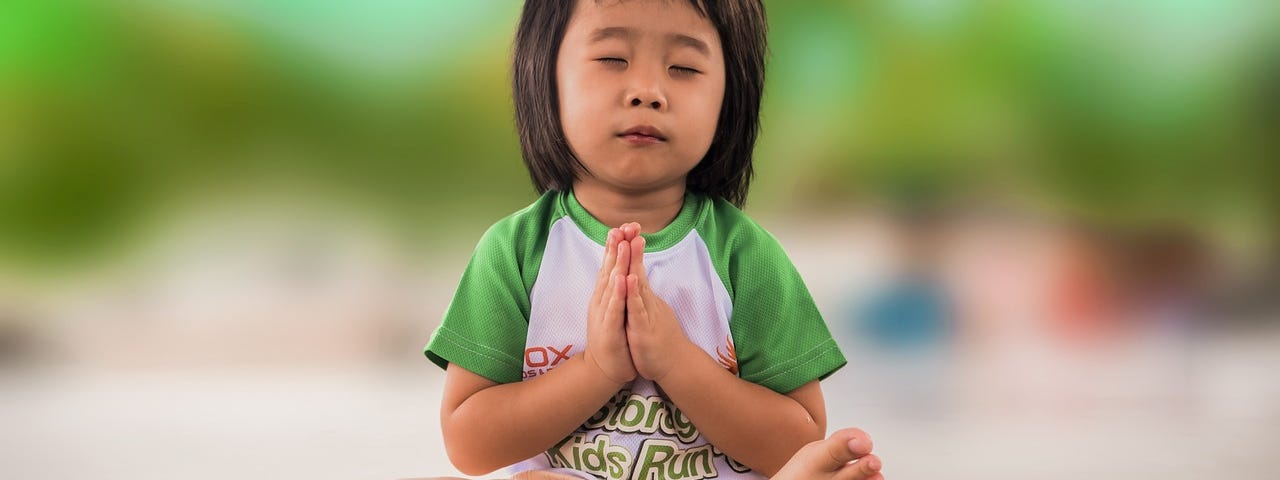 Young girl sitting in lotus position with hands folded in prayer, eyes closed.
