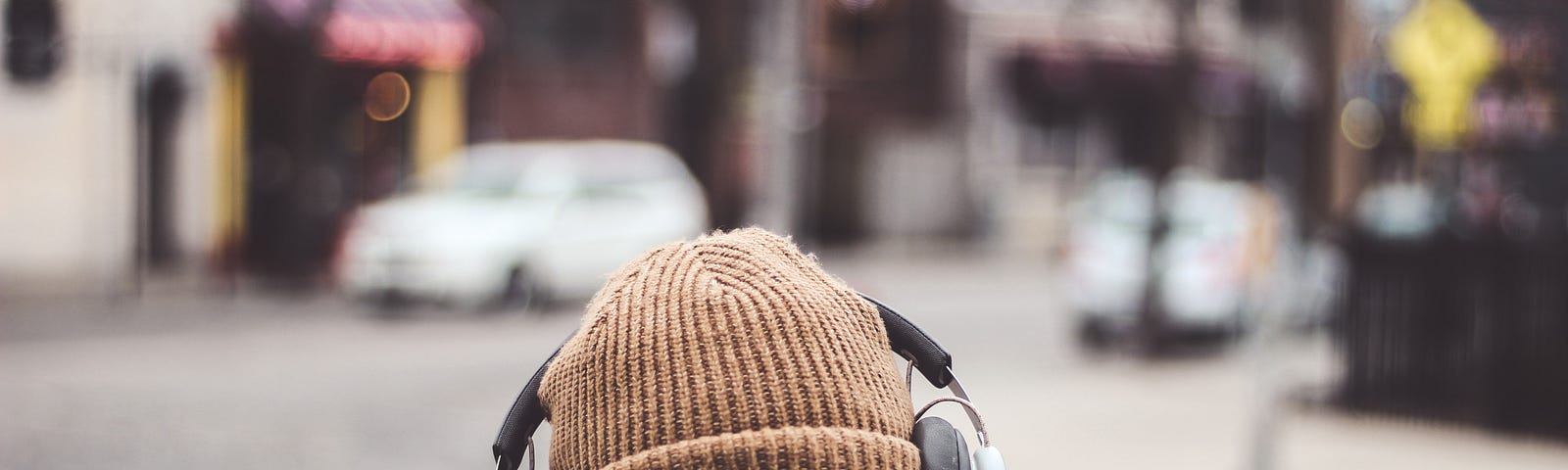 The photo captures the back of a person wearing a knit cap and over-the-ear headphones as they walk down the street, which is out of focus in the background.