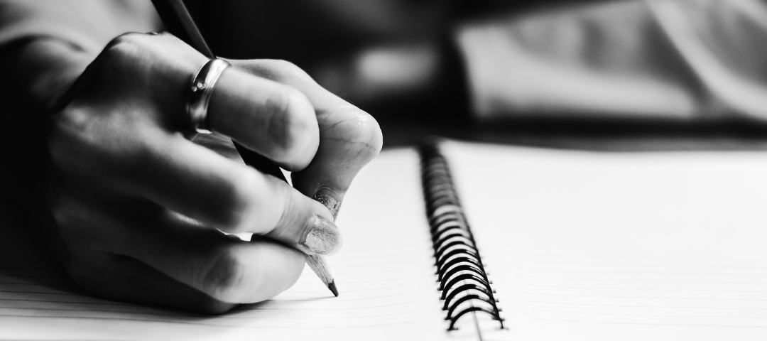 a person’s hand holding a pencil and writing in a spiral-bound notebook