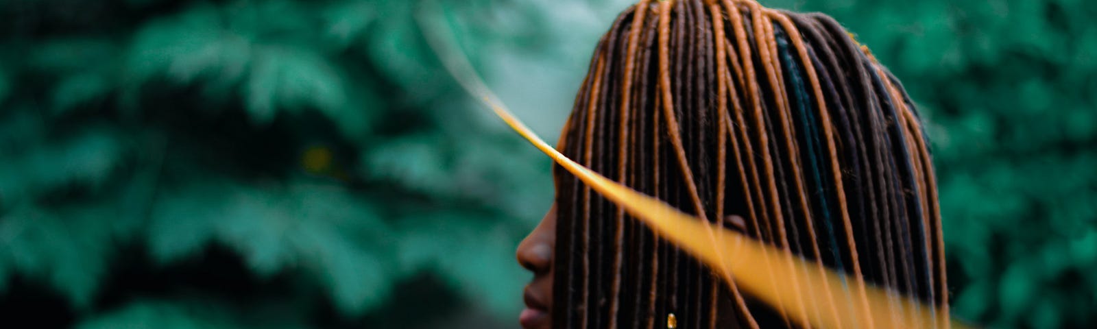 African woman side-on, face hidden with black and brown braids with a backdrop of green leaves