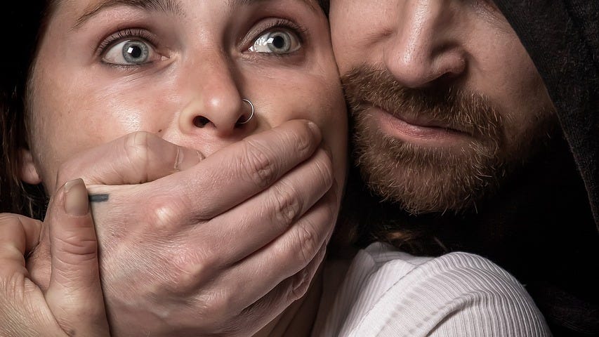 A woman in fear, caught from behind,  by a man woth his hand over her mouth.