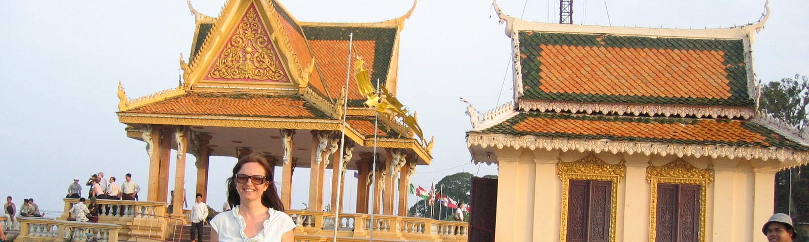 Lisa in front of a pagoda on the riverside in Phnom Penh, Cambodia. She is wearing a light blue sleeveless shirt and black trousers. She has long brown hair and is wearing and sun glasses. In the middle distance a Cambodian man cycles past smiling at the camera. In the background people sit and stand looking out over the river.