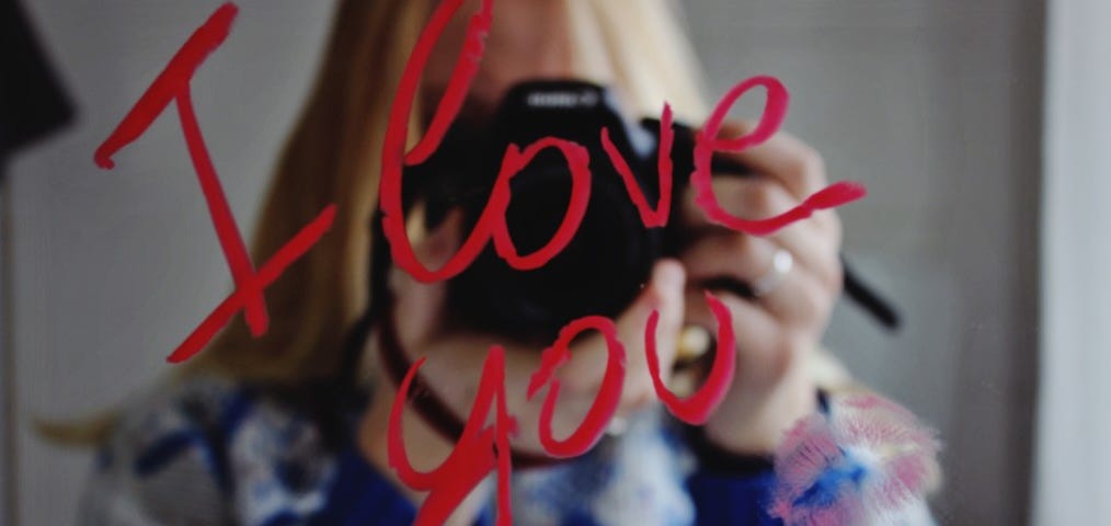 Red text reading “I love you” is written on a mirror. A woman photographs it in the background.