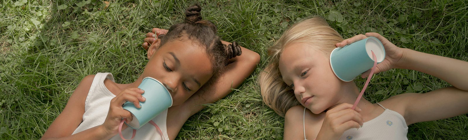 Two young girls laying on grass play telephone with cups and string.