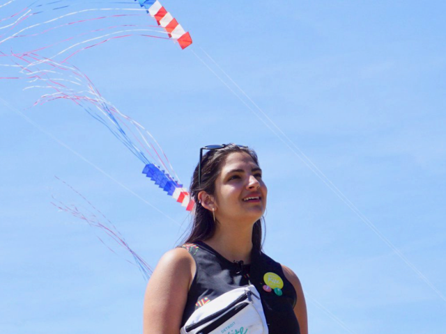 Margo Dalal, a young woman, looks to the sky with a smile during the Detroit Kite Festival. Kites fly in the background.