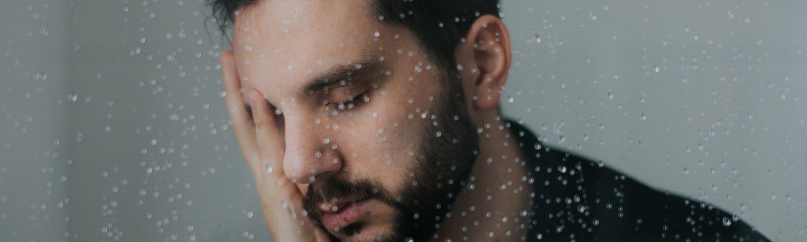 An anxious man rests his head in his hands behind a rain-spattered pane of glass.