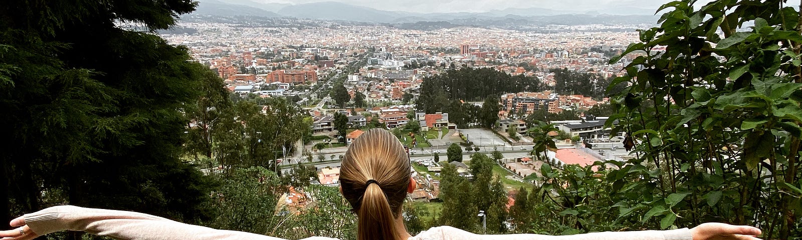 Woman standing with arms spread. The city of Cuenca is in the distance nestled amongst lush green trees and mountains.