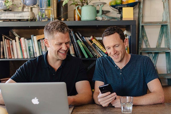 Two men sitting next to each other with laptop and cell phone.