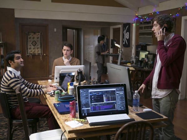 A screenshot from HBO’s “Silicon Valley” television show where three middle-aged men are gathered around a rectangular table full of computers (two people are seated, one is standing on his phone). The table is messy and filled with water bottles, disposable coffee containers, and office knick knacks.