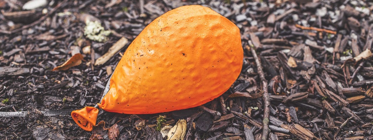 A deflated orange balloon laying alone on damp woodchip ground