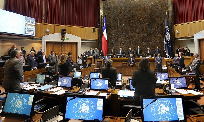 Image of Chilean senate hall: many desktops are on long wooden desks that form a semi-circle infront of a podium which features men in suits and the chilean flag.