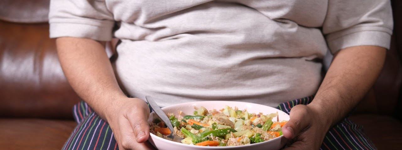 An obese person (whose face is cut off in the photo) sits on a brown leather sofa and holds a bowl of what might be some type of stir fry or pasta filled with vegetables.