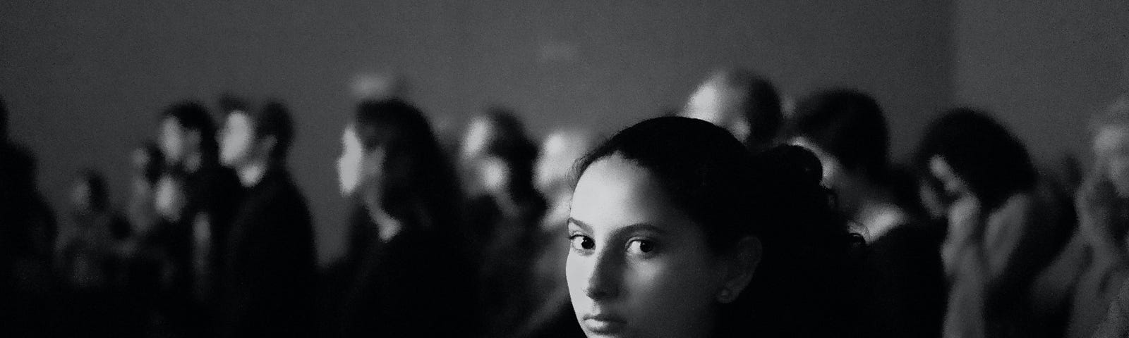 A black and white photo with a young woman gazing directly at the camera with people beyond looking in a different direction.