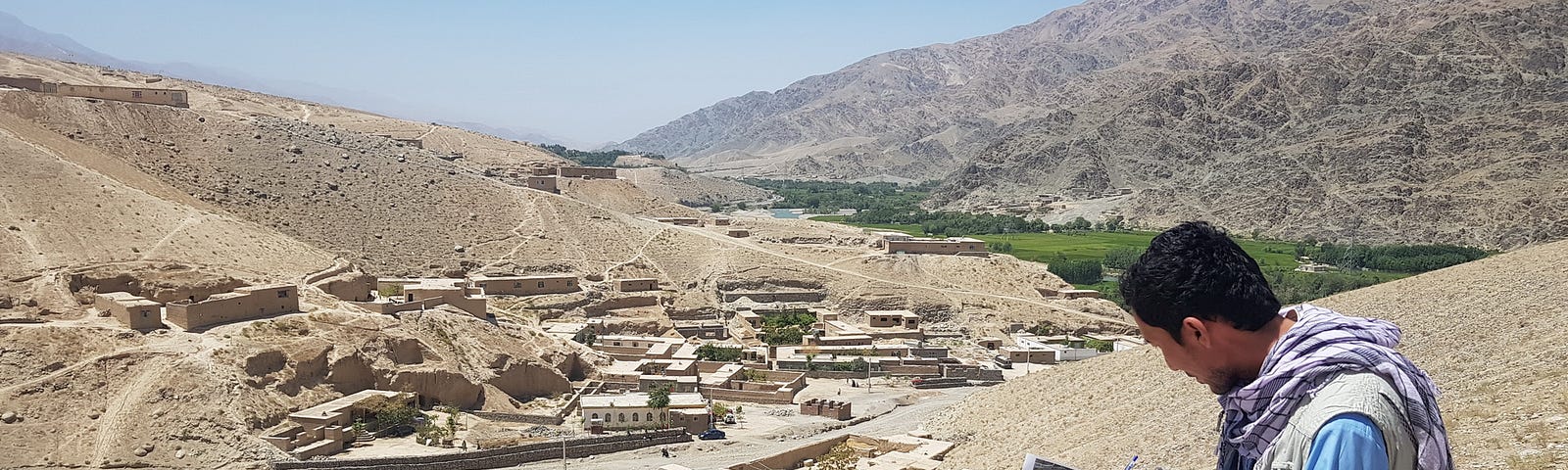 A man crouches and records notes on a map. In the background, there is a village at the foothills of two mountains.