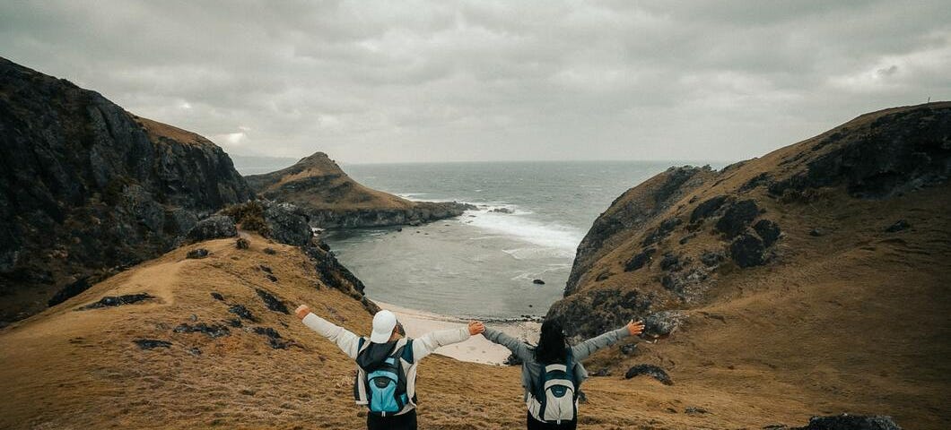 Two travelers stand at the top of a cliff and overlook the sea with their arms outstretched.
