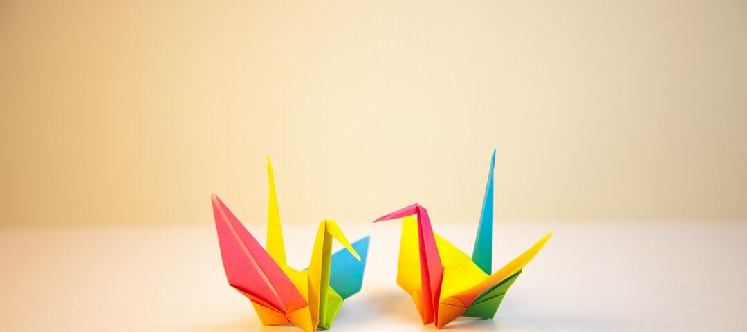 Two origami cranes made out of brightly covered paper sit on a table facing each other.