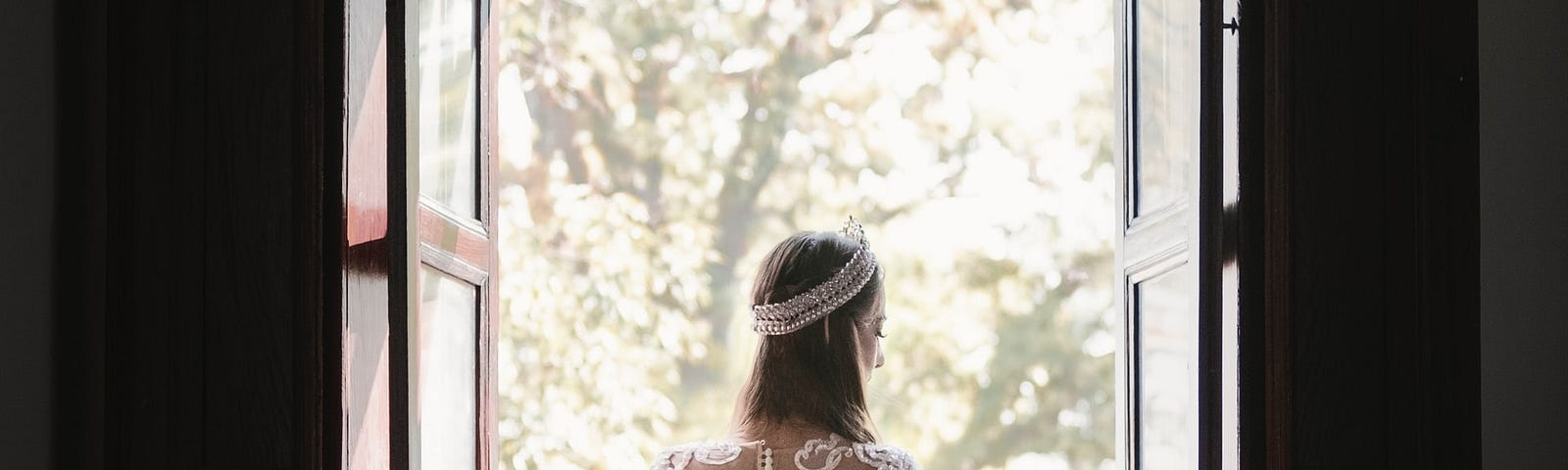 Young woman opening a door on to a garden. She is dressed in a crown and a lace dress and gloves like it’s her fantasy wedding day.