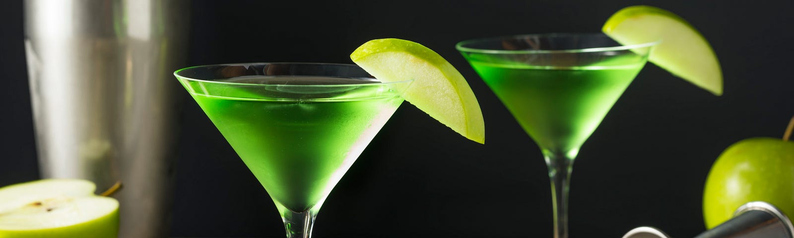 Two tempting, green apple-tinis on a rough wood bar, one of a silver serving tray. A shaker, a jigger, and green apples appear with the cocktails, against a black background. Depositphotos.