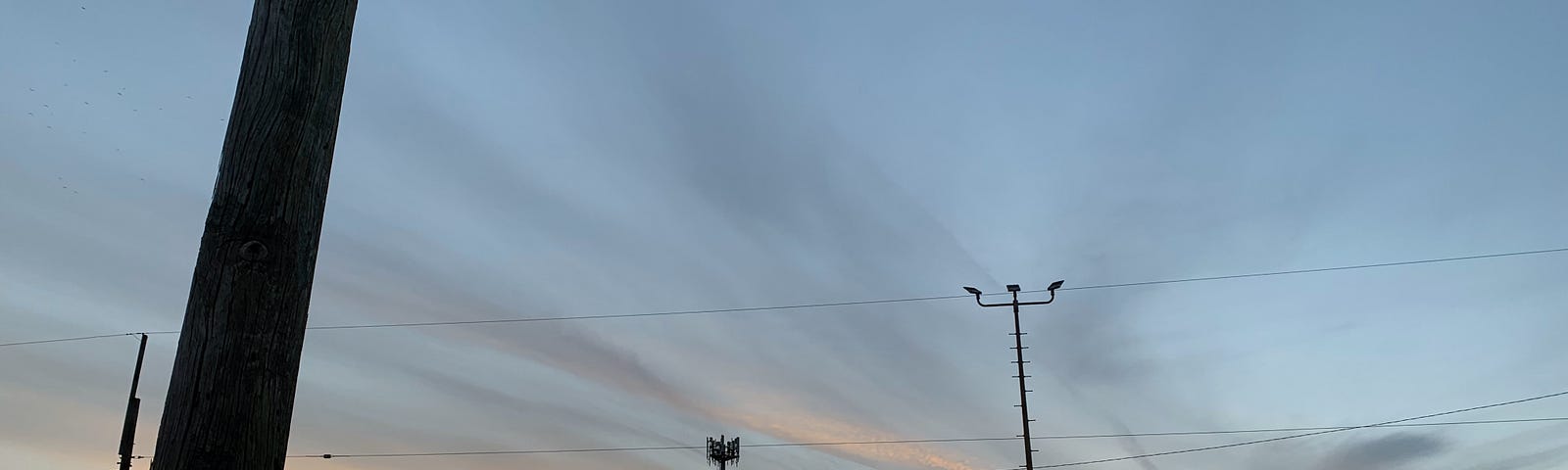 A sunset of pink and grey at the horizon in a light blue sky. a shadow of a water tower, electrical poles / wires, & a street