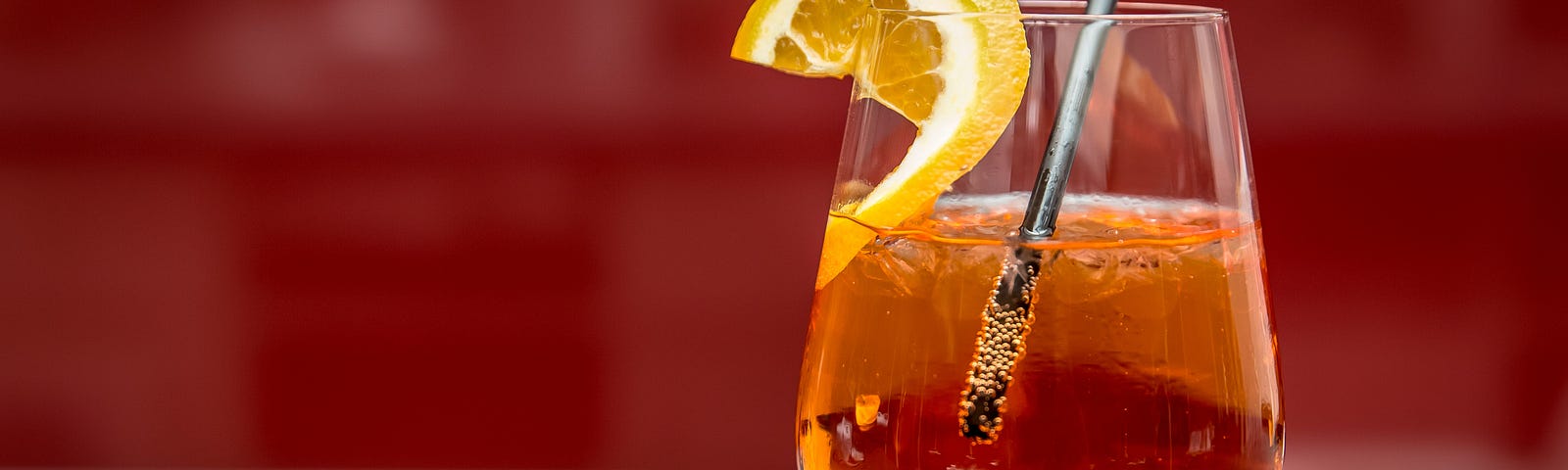 A stemmed glass with an amber-coloured cocktail over ice sits in front of a blurred grey and cool red background. The glass has a short black straw and is garnished with a slice of lemon on the rim.