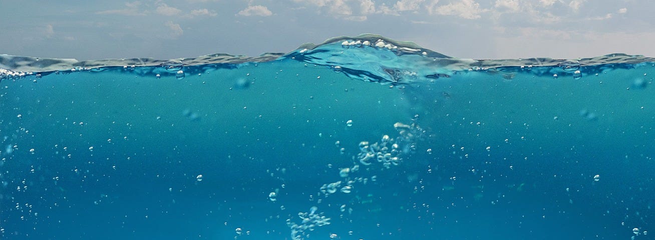 Cross-sectional view of cloudy, bright blue skies above turquoise blue waters with light ripples and air bubbles dotting across the water surface and the sea bed visible underneath.