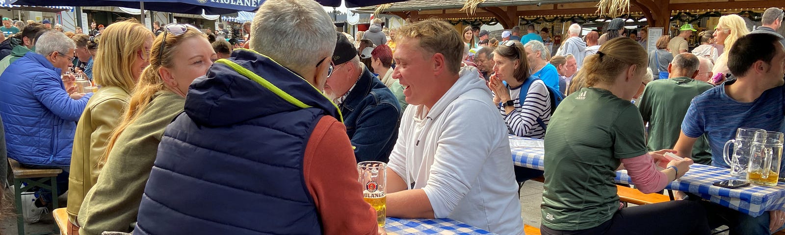 A large group of people sit on benches at tables covered in blue and white checked tablecloths. Some have large steins of beer. And there are blue umbrellas over some of the tables.