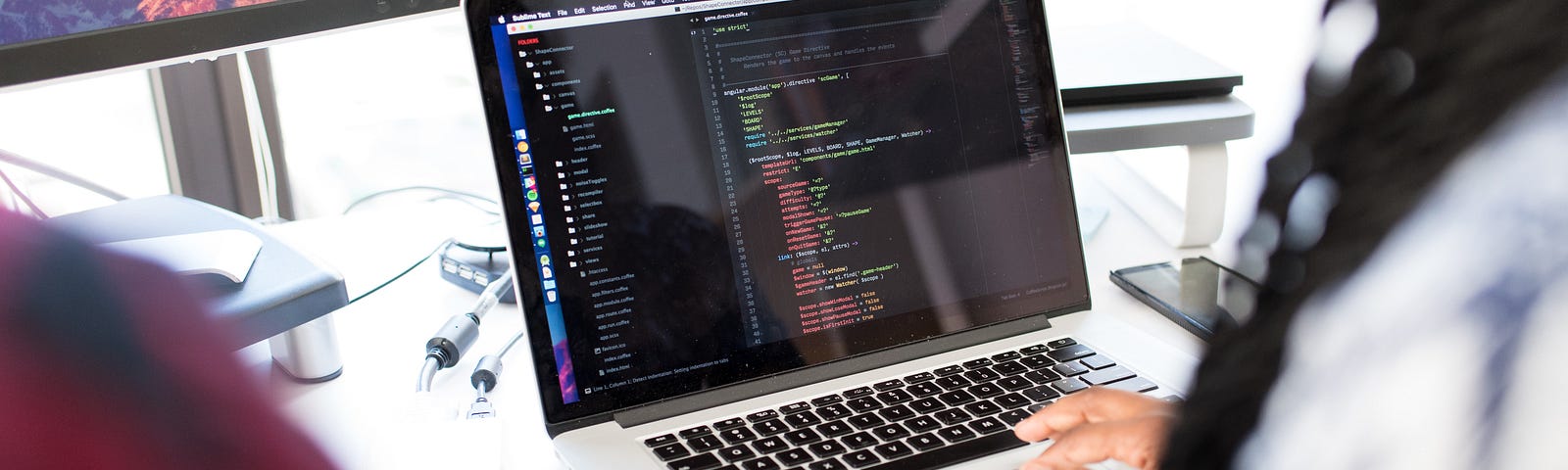 Dark skinned woman typing code on a laptop. We can also see the shoulder of someone else just outside of the frame of the photo.