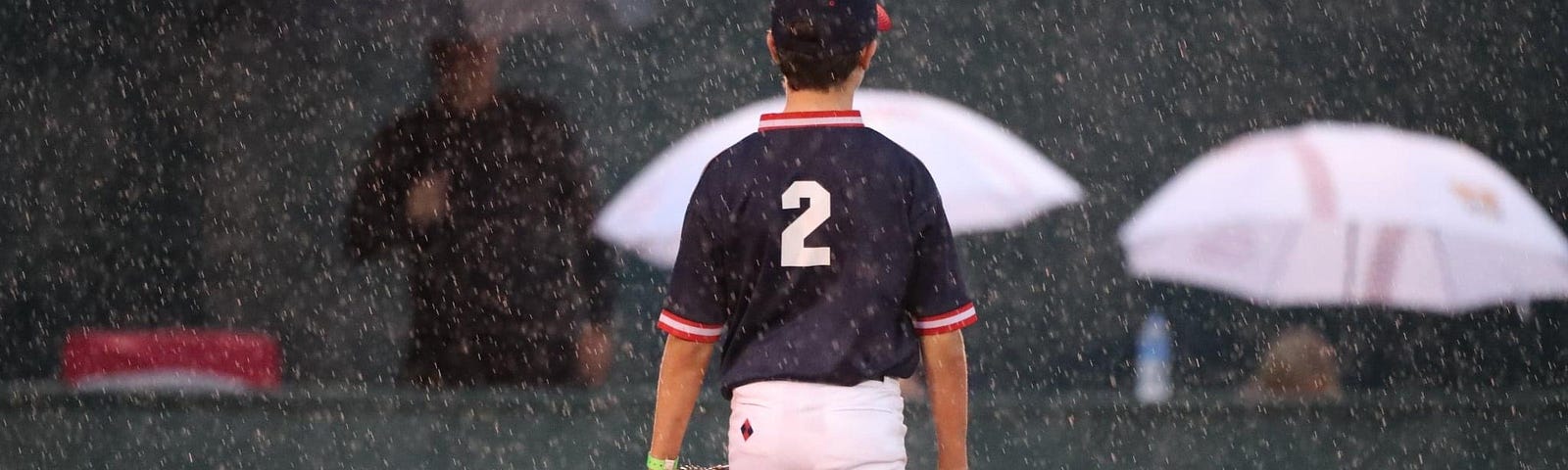 A 12-year-old baseball player stands alone on the infield grass in the rain.