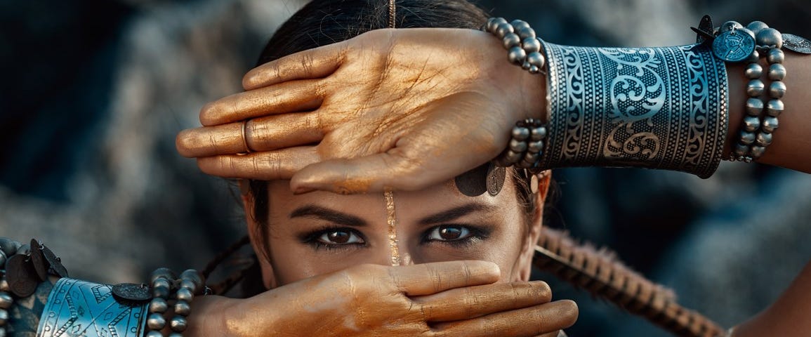 A female tribal warrior stands with her hands outstretched on her face, leaving only her eyes uncovered.