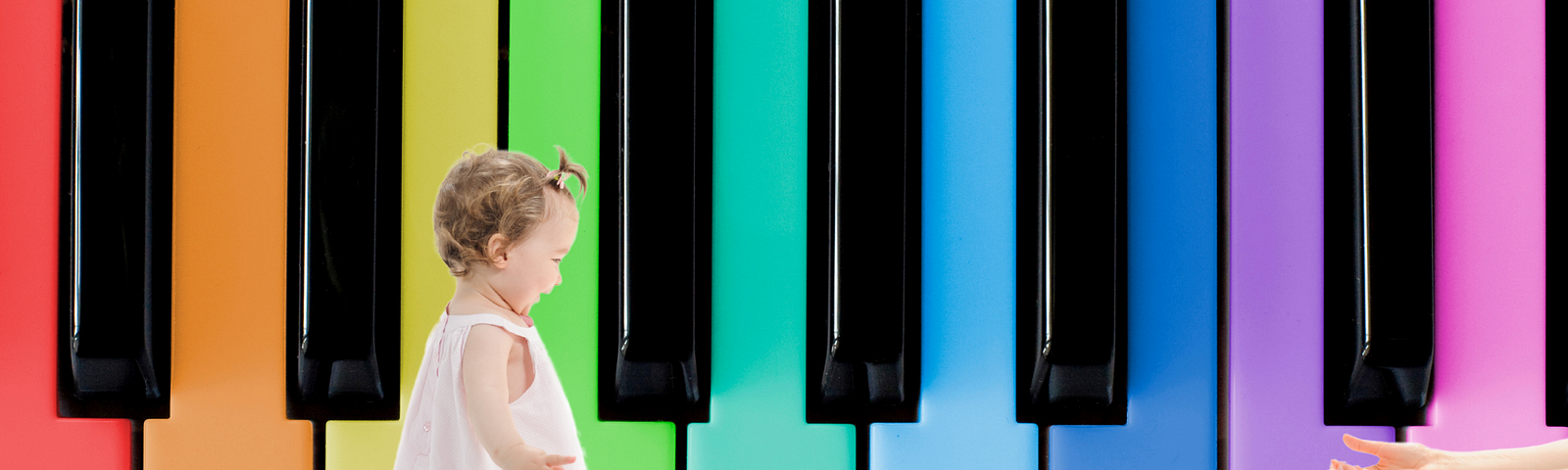 You see the keys of a piano where the white keys are replaced by colorful keys (orange, yellow, green, turquoise, blue, purple, pink). A toddler in a light pink dress with a happy expression on her face seems to be taking her first steps on the piano keys, towards two hands inviting her to get there.