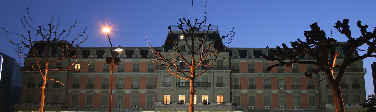 Evening photo of the Palais Wilson with darkening sky and blurred vehicle lights in the foreground.