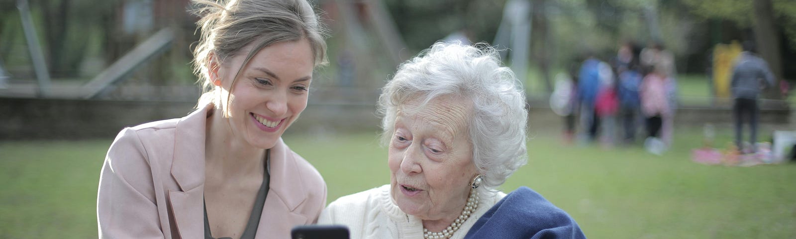 A woman in her 30s helps an elderly woman with something on her phone.