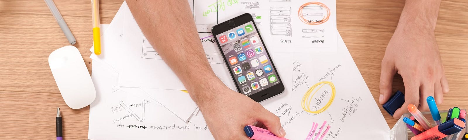 A person marking notes on various business strategy documents with colorful pens, with a smartphone displaying multiple apps and an iMac on a wooden desk.