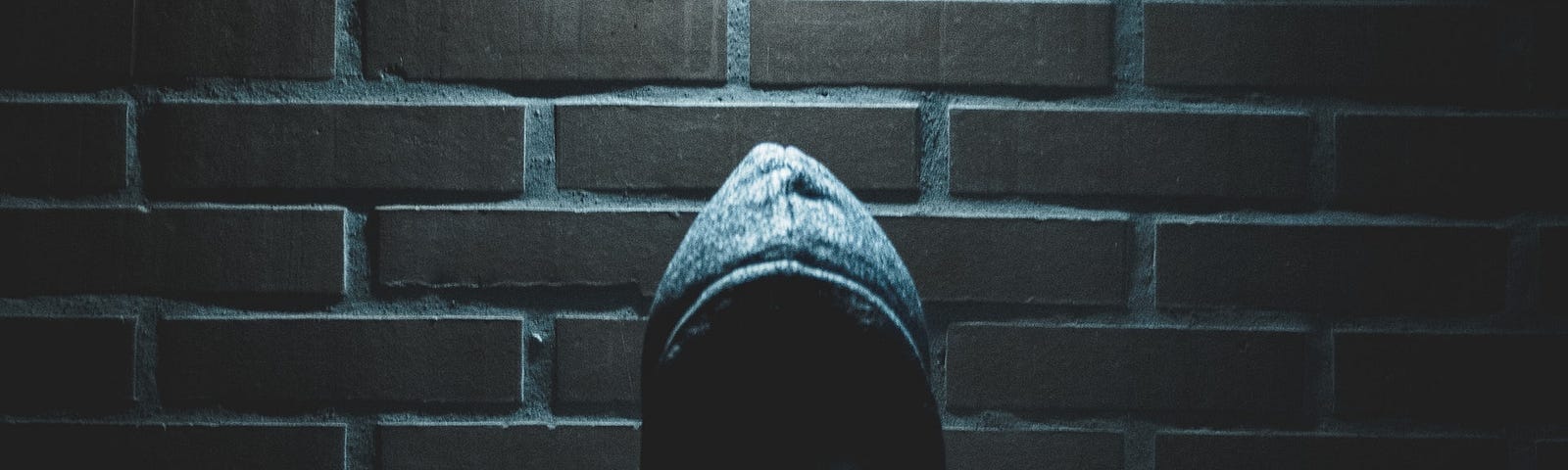 Image of a hooded man, face not visible, stood against a brick wall, beneath a light