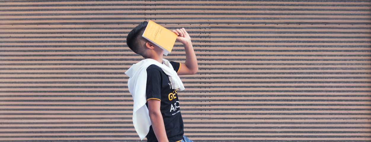 Man walking with one leg extended in front of him, holding a yellow book open against his face