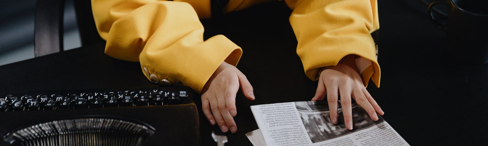 A child’s hands, in a yellow jacket, adjust a newspaper with a typewriter nearby