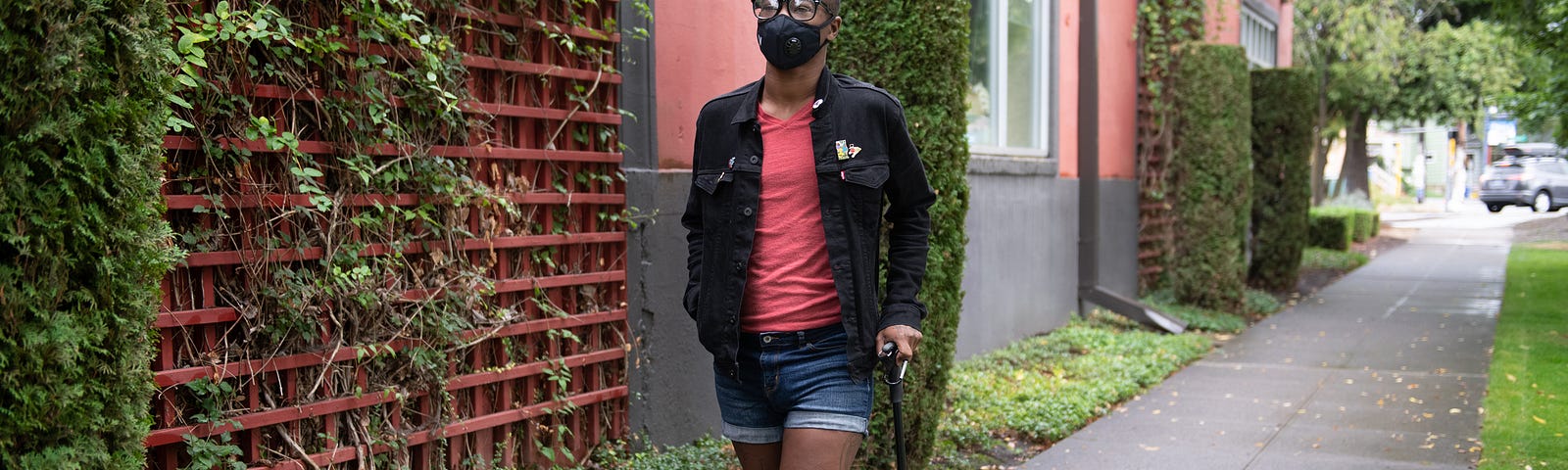 A Black non-binary person with a filtering face mask walks down a neighborhood street with one hand in their pocket