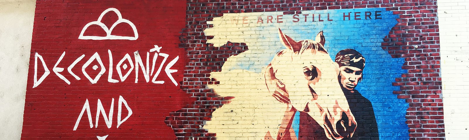 A mural painted on a brick wall depicting a man leading a white horse. The phrase “we are still here” is written above the mural, while the phrase “Decolonize and Chill” is written next to it.
