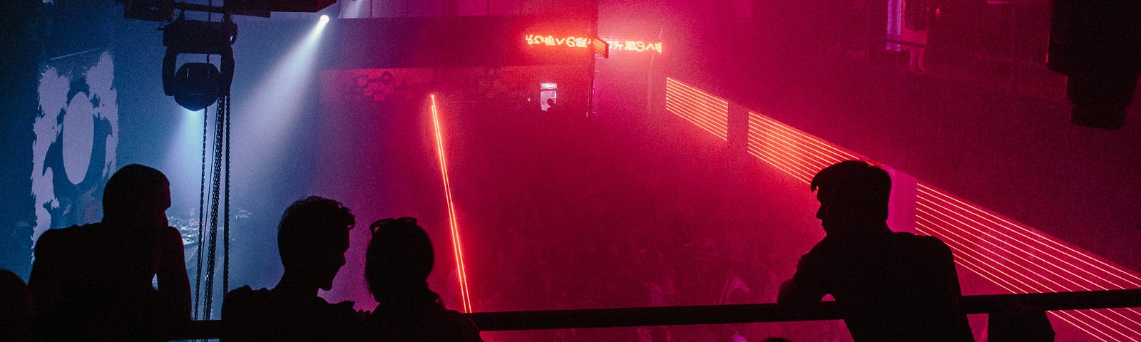 Silhouettes of men leaning against a railing overlooking a dancefloor bathed in pink light.