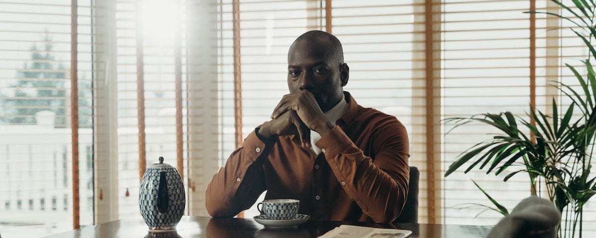 A Black man, dressed smartly but casually, sits at a boardroom table, in front of a newspaper, a pot of coffee, and a coffee cup.