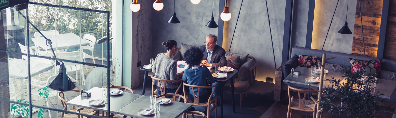 A photograph of the interior of a modern restaurant. Three people are sat at a table, having a business meeting. Two people have their backs to the camera; one man is facing them across the table.