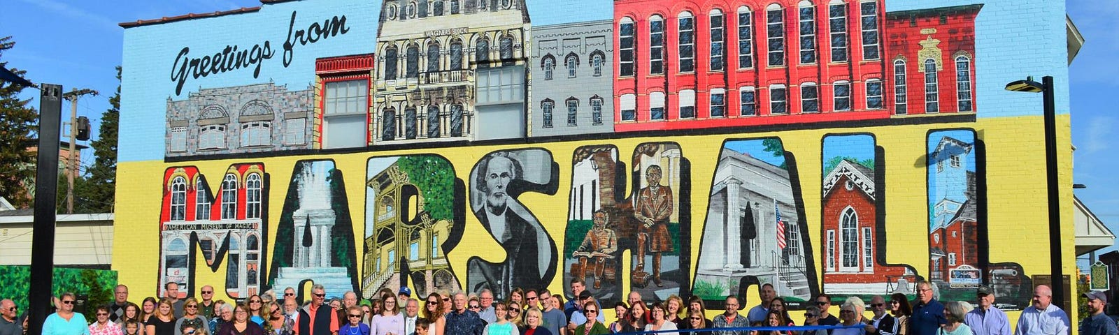 The ribbon cutting ceremony for Grand Street Park in Marshall, Michigan. Hundreds of community members stand behind a blue ribbon in the park in front of a large mural with the words, “Greetings from Marshall.”